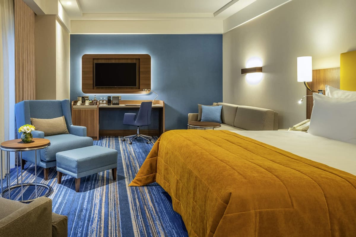 A hotel room with a large bed, a desk with a chair, a sofa, and a small table with a vase.