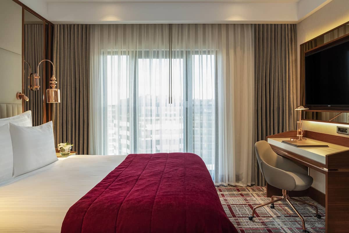 A hotel room with a large bed, a desk with a chair, a television, and a city view through the window.