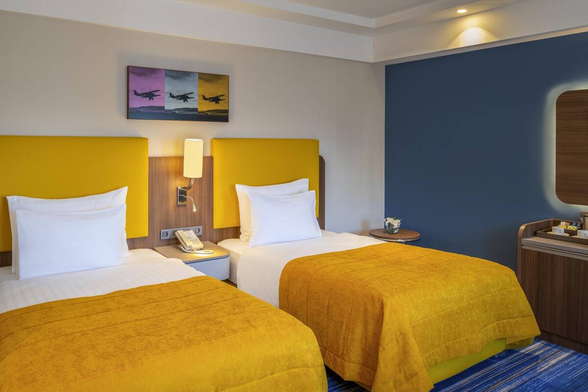 A hotel room with two beds, a bedside table with a lamp, a phone, and a painting on the wall.