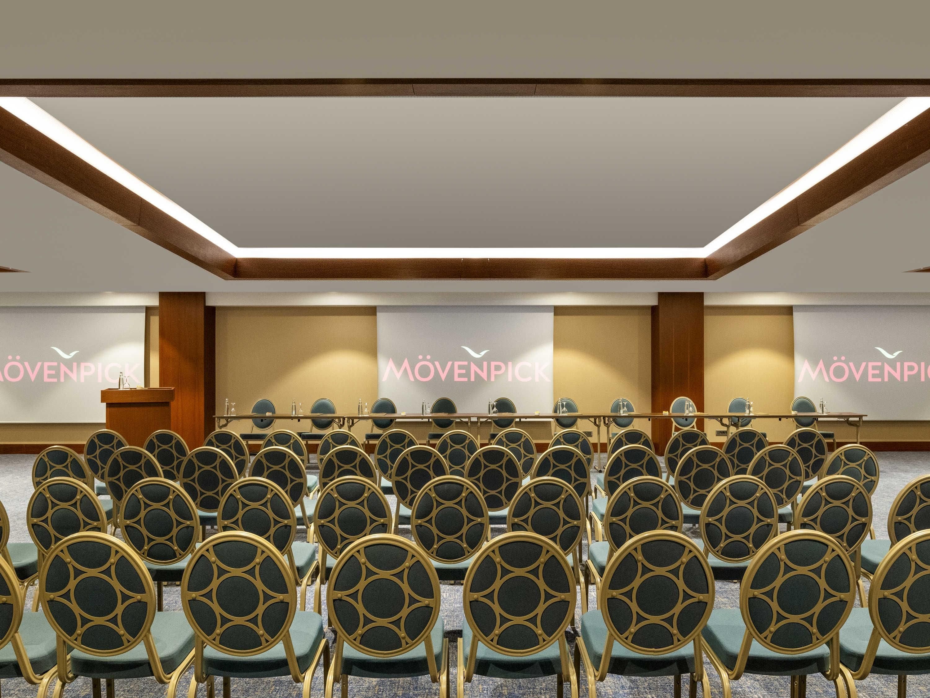 Un salón de conferencias con filas de sillas, un atril y una pantalla de proyecciones con el texto 'MÖVENPICK'.