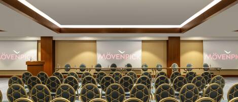 Un salón de conferencias con filas de sillas, un atril y una pantalla de proyecciones con el texto 'MÖVENPICK'.