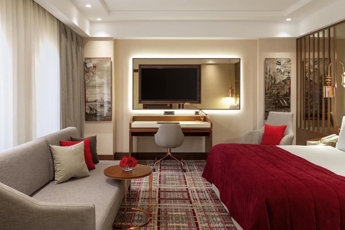 A hotel room with a sofa, a bed, a desk with a TV, a small table with flowers, and artwork on the walls.