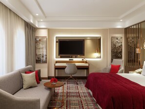 A hotel room with a sofa, a bed, a desk with a TV, a small table with flowers, and artwork on the walls.