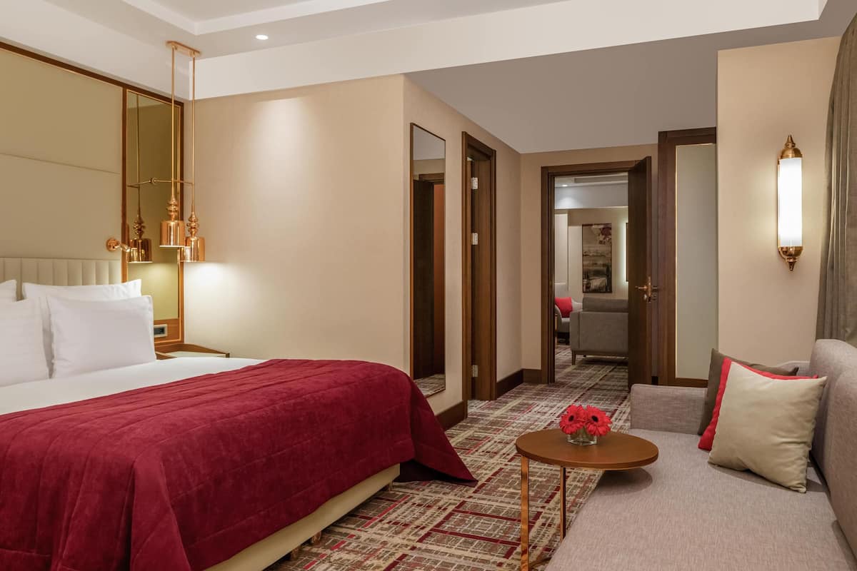 A hotel room with a large bed, a sofa, a small table with a vase of flowers, and a mirror.