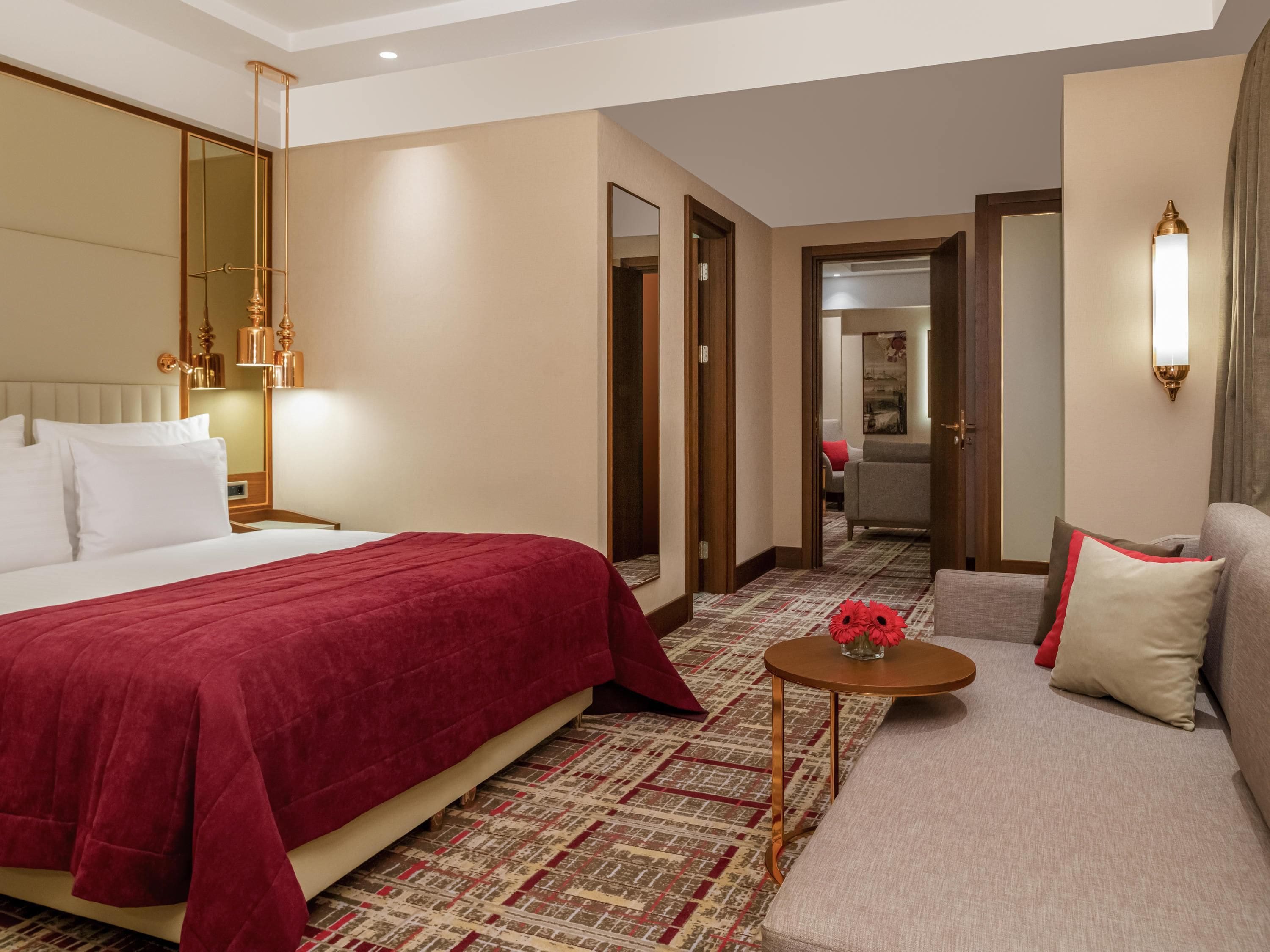 A hotel room with a large bed, a sofa, a small table with a vase of flowers, and a mirror.