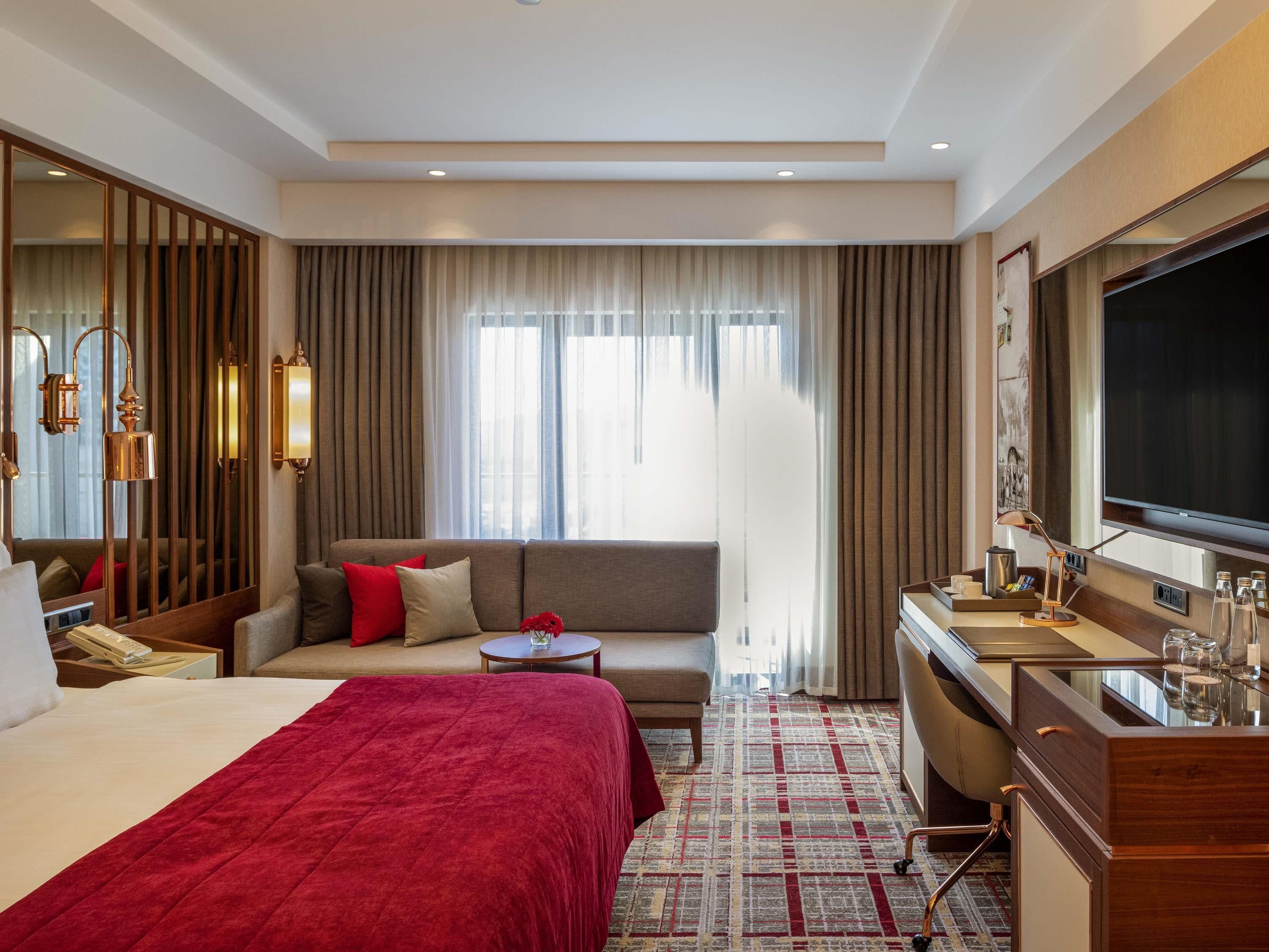Een hotelkamer met een groot bed, een bank, een bureau met een televisie en een groot raam met gordijnen.