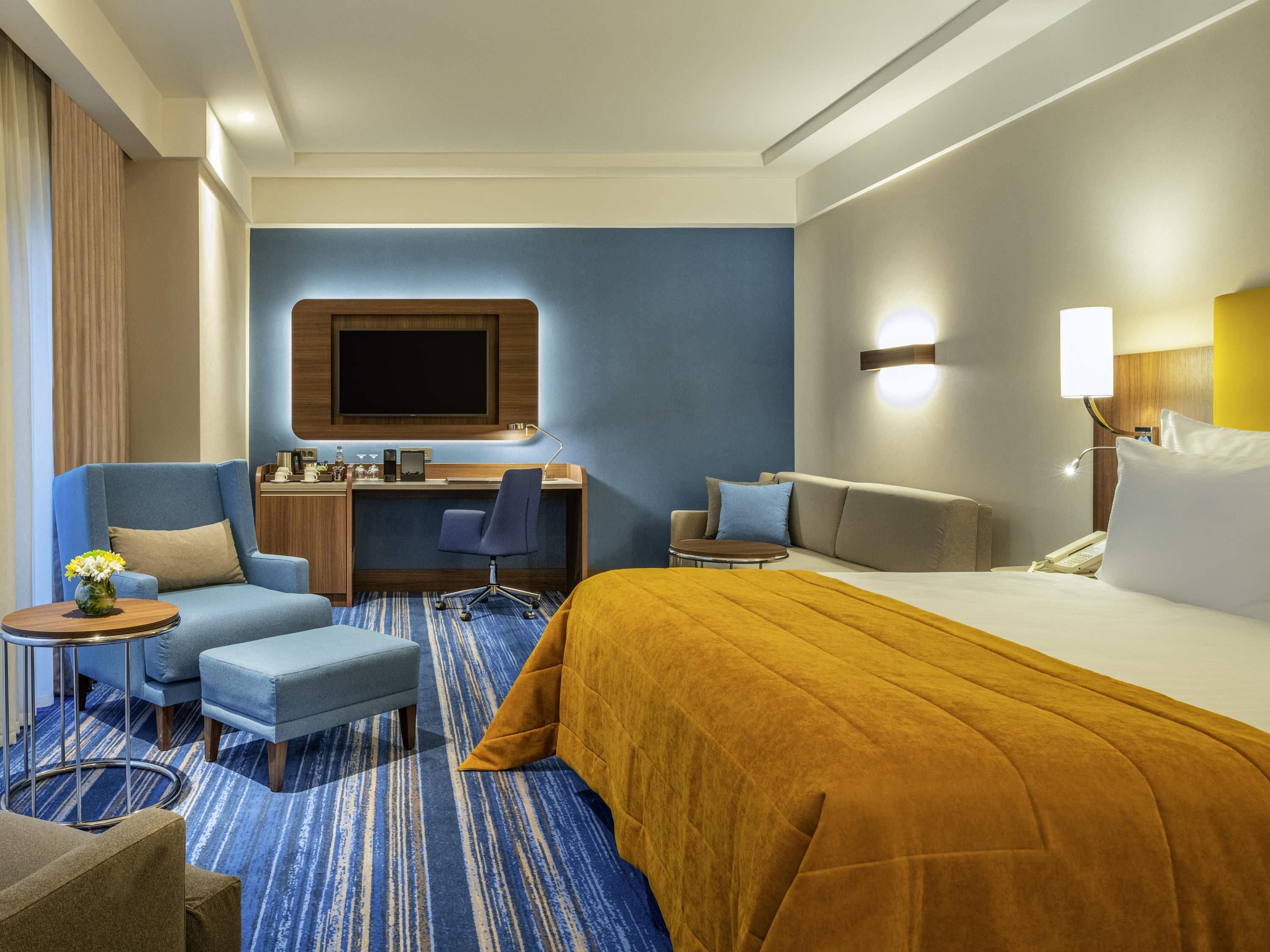 A hotel room with a large bed, a desk with a chair, a sofa, and a small table with a vase.
