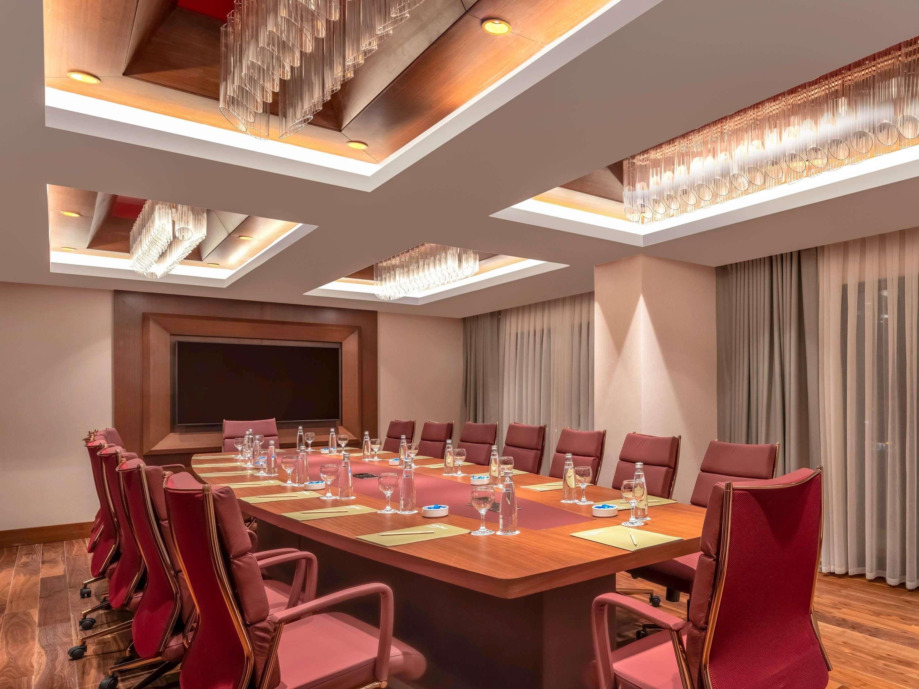 Un salón de conferencias con una larga mesa, sillas rojas y una pantalla grande.