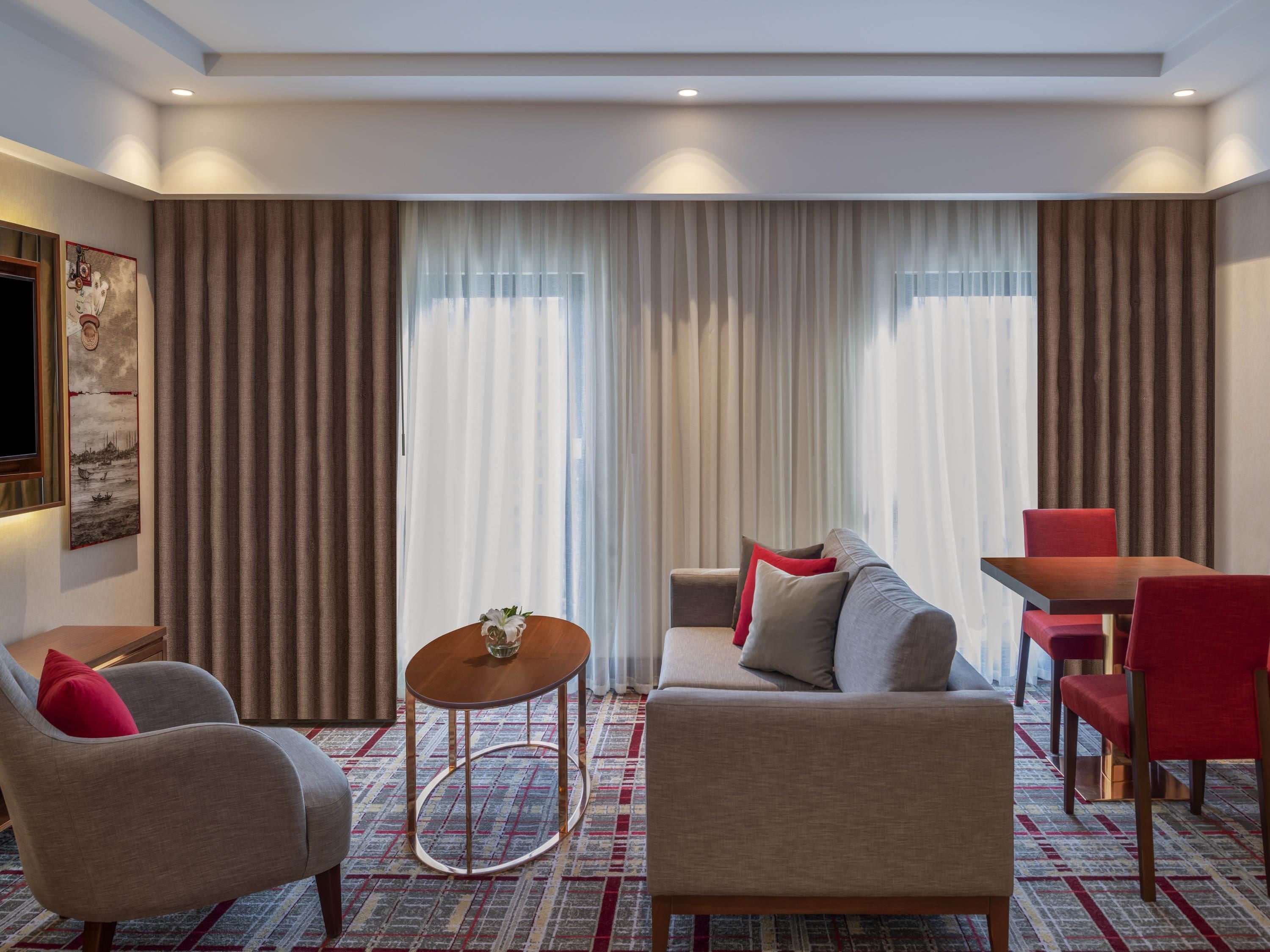 A hotel room with a sofa, armchairs, a small round table, and a dining area with a table and chairs.