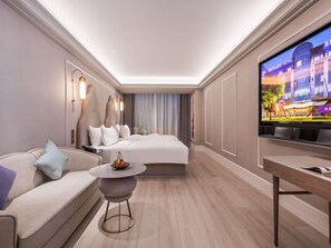 A modern hotel room with a large bed, a sofa, a small round table, and a TV displaying a hotel image.