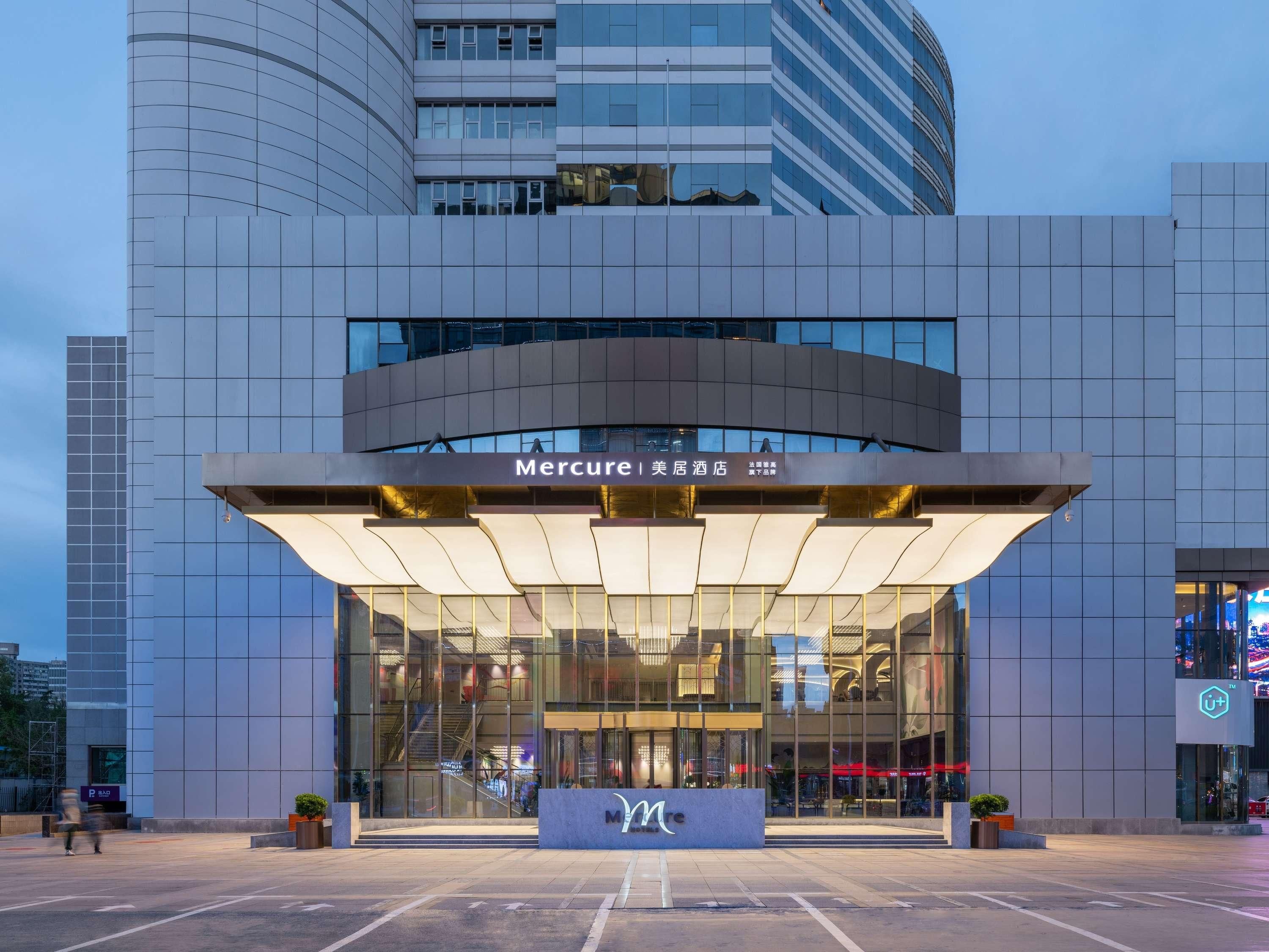 Une entrée d’hôtel moderne avec une façade en verre, une enseigne « Mercure » bien visible et une vue dégagée sur l’environnement urbain.