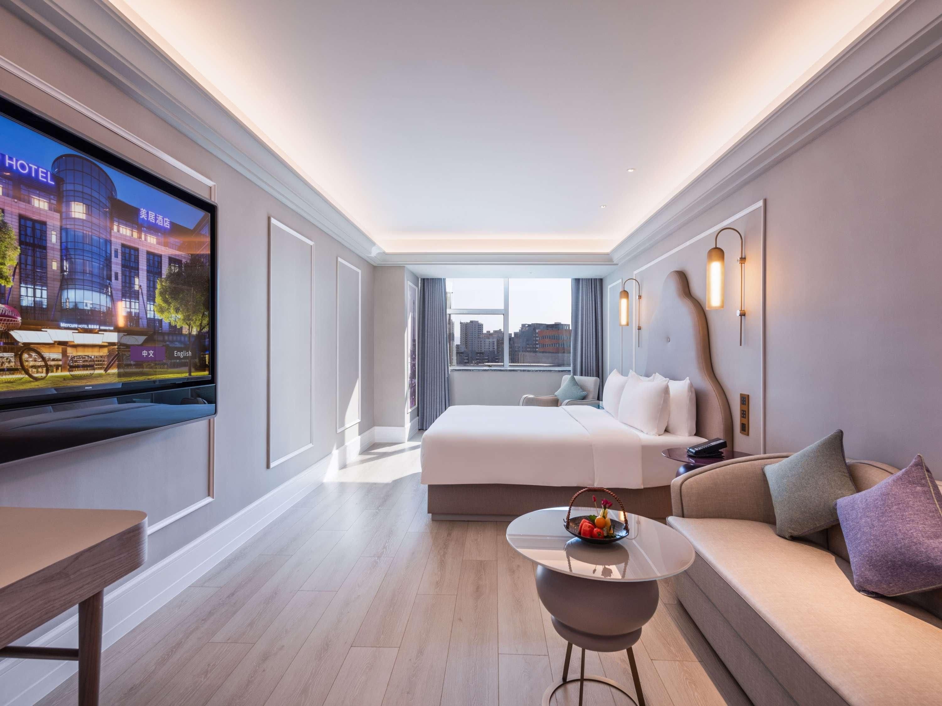 A modern hotel room with a large bed, a small round table with a fruit basket, a sofa, and a TV displaying a hotel sign.