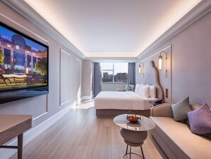 A modern hotel room with a large bed, a small round table with a fruit basket, a sofa, and a TV displaying a hotel sign. - Mercure Shijiazhuang People Square (Shijiazhuang)