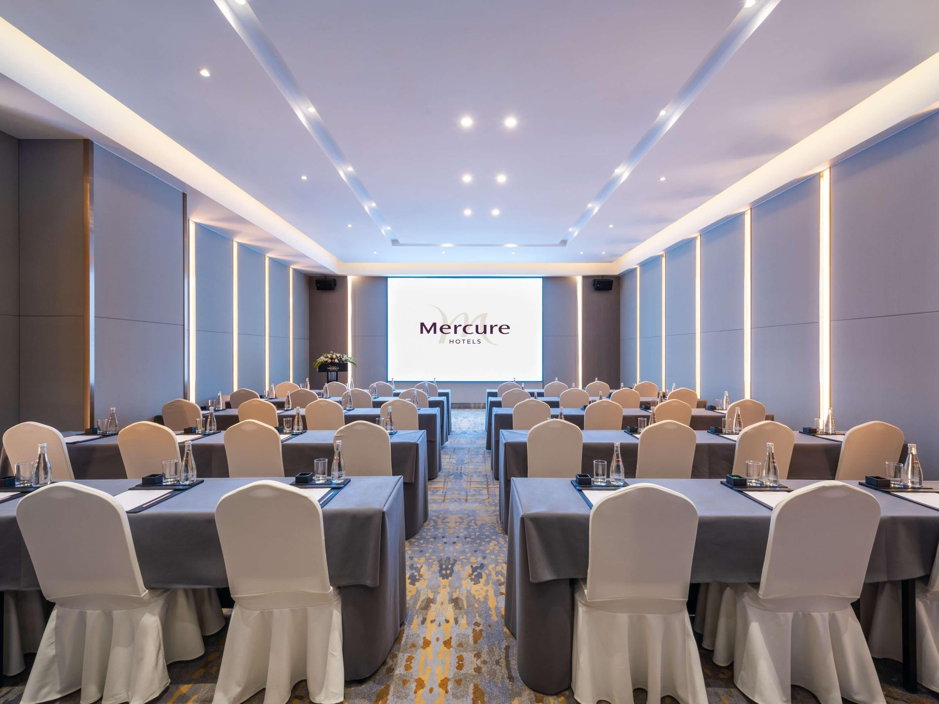 Une salle de conférence dotée de plusieurs tables et chaises, d’un grand écran affichant le logo « Mercure Hotels », et d’un sol recouvert de moquette.