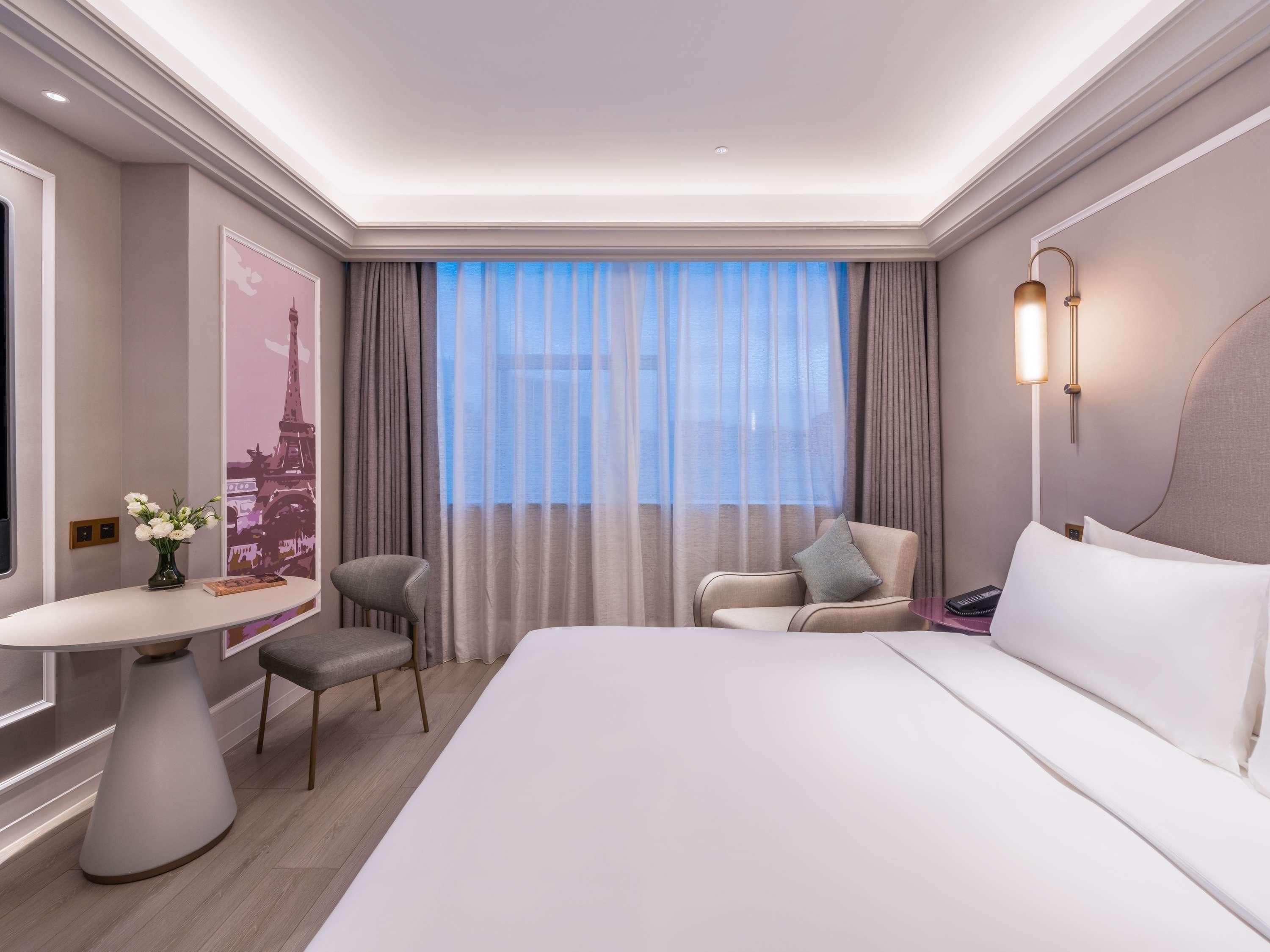 Une chambre d’hôtel moderne avec un grand lit, une table ronde ornée d’un vase de fleurs, deux fauteuils, une grande fenêtre avec des rideaux légers et une lampe fixée au mur.