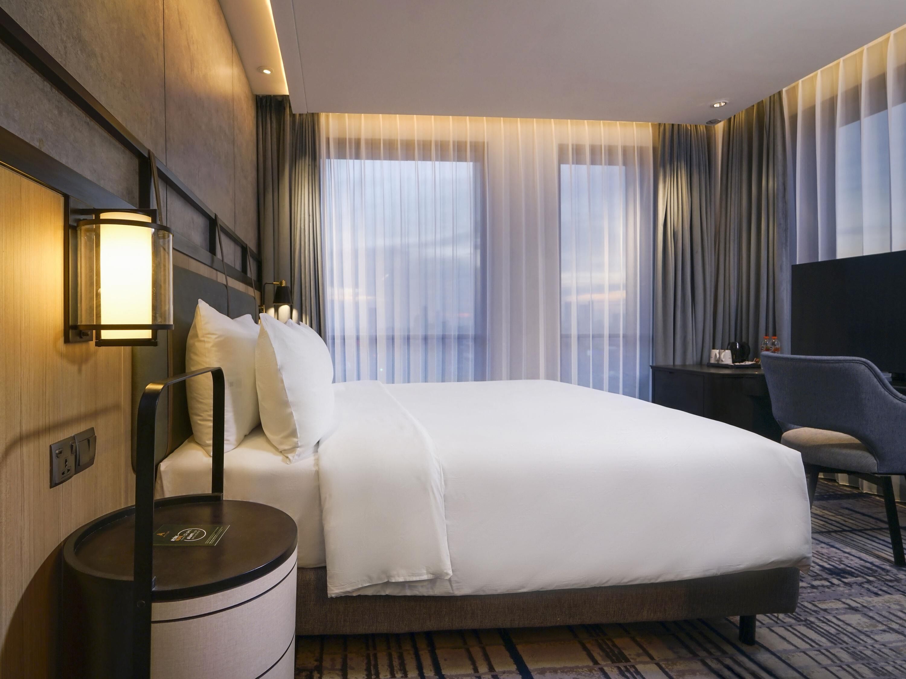 A hotel room with a large bed, a bedside table, a chair, a TV, and a window with curtains.
