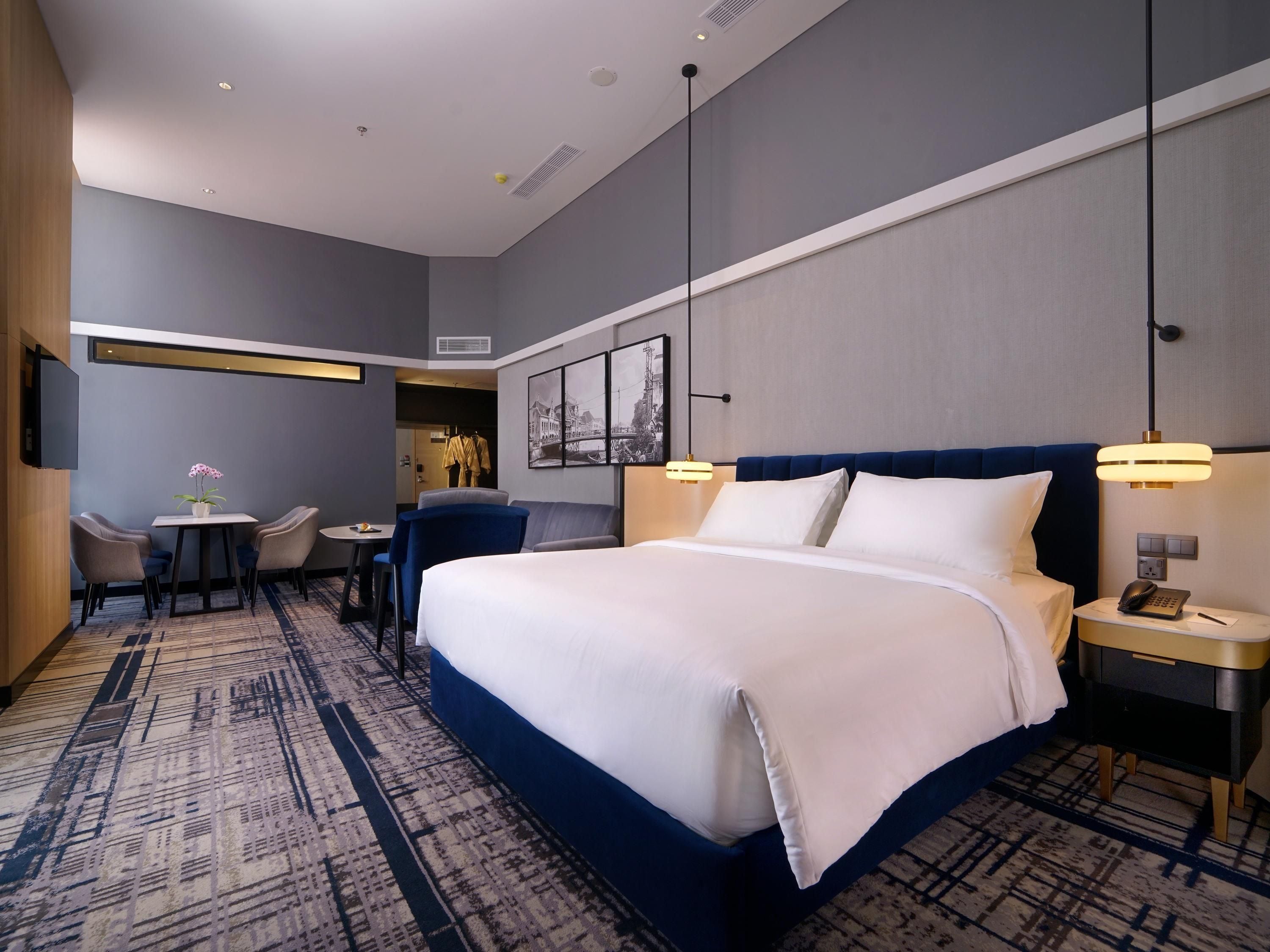 A hotel room with a large bed, a desk with a chair, a small table with a vase, and a wall-mounted TV.