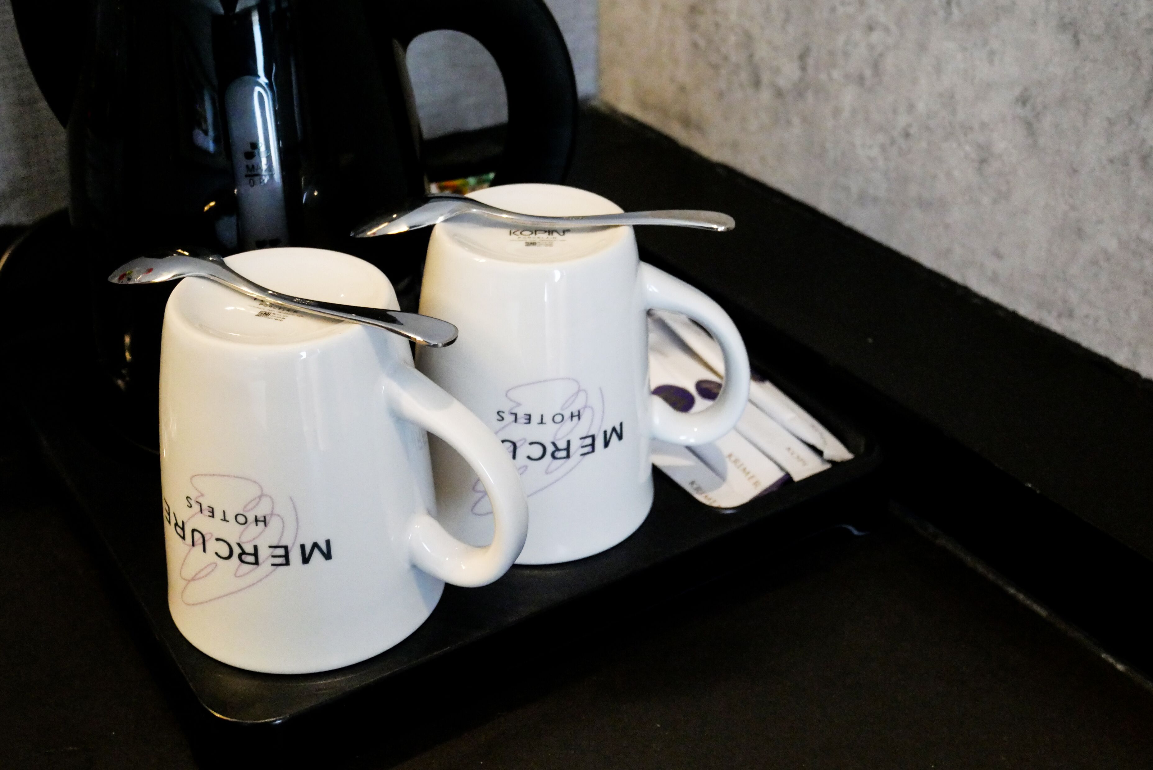 Two white mugs with "Mercury Hotels" text and a spoon on a black tray.