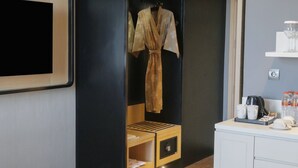 Quarto de hotel com guarda-roupa preto, roupão pendurado, pequeno armário de madeira e balcão branco com chávenas e garrafas.