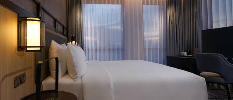 A hotel room with a bed, bedside table, lamp, chair, TV, and curtains.