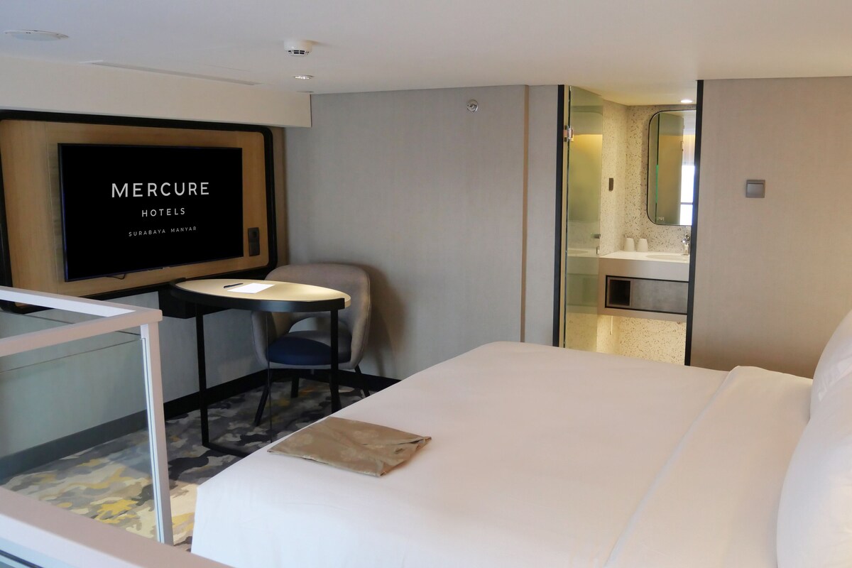A hotel room with a bed, a desk with a chair, a television displaying 'MERCURE HOTELS SURABAYA MANYARA', a bathroom with a mirror, and a bedside table with a lamp.