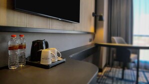 Quarto de hotel com televisão de tela plana na parede, cafeteira, duas garrafas de água e uma bandeja com xícara e controle remoto.