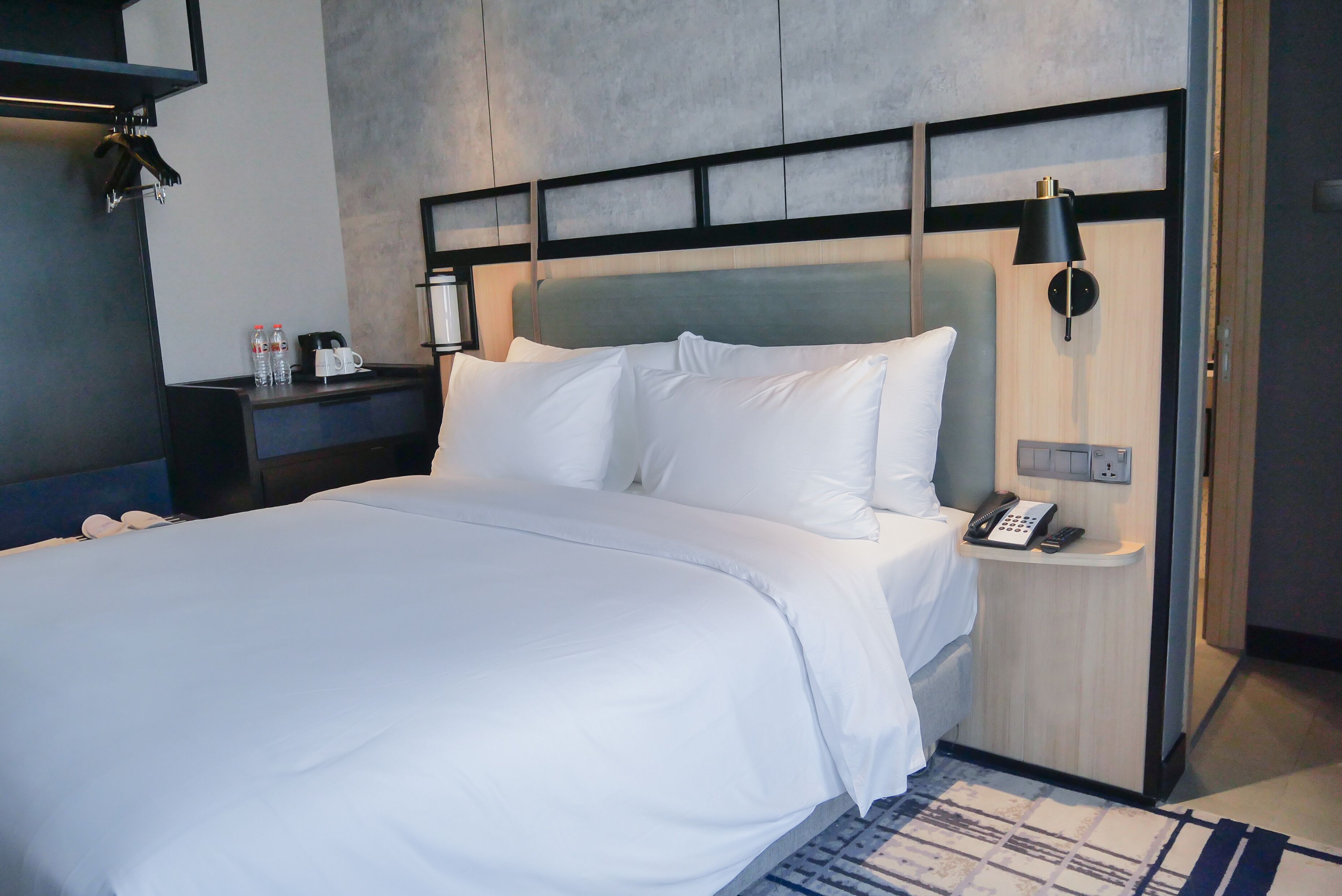 A hotel room with a large bed, a bedside table with a phone and a lamp, and a wall-mounted shelf with bottles.