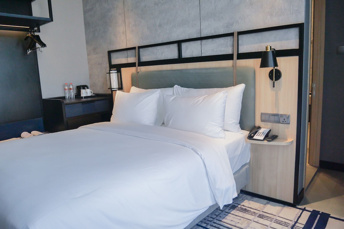 A hotel room with a large bed, a bedside table with a phone and a lamp, and a wall-mounted shelf with bottles.