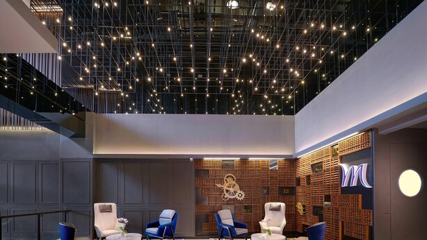 Um lobby moderno com design geométrico no teto, área de estar com cadeiras azuis e brancas, e uma parede com painéis de madeira e a placa 'YM'.