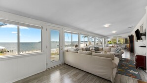 Amplia sala de estar con un sofá beige grande, piso de madera y vista al océano a través de ventanas amplias.