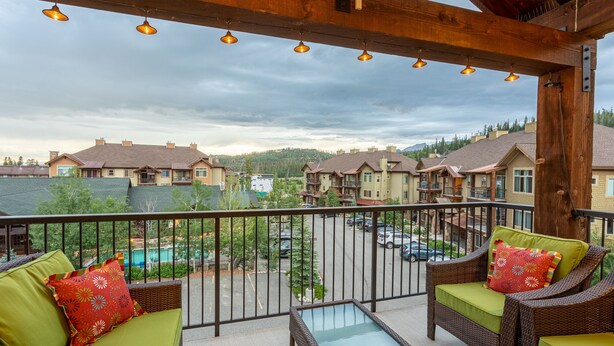 A balcony with wicker furniture and a glass table, overlooking a parking area and buildings.