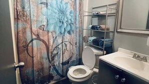 Ein Badezimmer mit Duschvorhang mit großem Blumenmuster, Toilette, Waschbecken mit Spiegel und Ablage über dem Waschbecken.