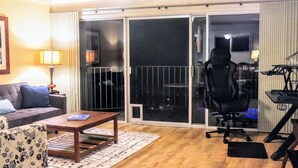 A living room with a sofa, a coffee table, a patterned rug, a gaming chair, and a desk with a computer. - Upgraded Makaha remote work and surf oceanfront condo (Waianae)