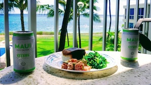 A table with a plate of food and two cans of 'Maui Hard Seltzer Citrus' in a room with a view of the ocean and palm trees. - Upgraded Makaha remote work and surf oceanfront condo (Waianae)