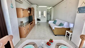 Un espace de vie compact comprenant un coin cuisine, un canapé, une table à manger pour deux personnes et une vue sur la pièce adjacente.