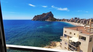 Une vue côtière avec une île rocheuse, une mer d’un bleu limpide et une plage bordée de bâtiments.
