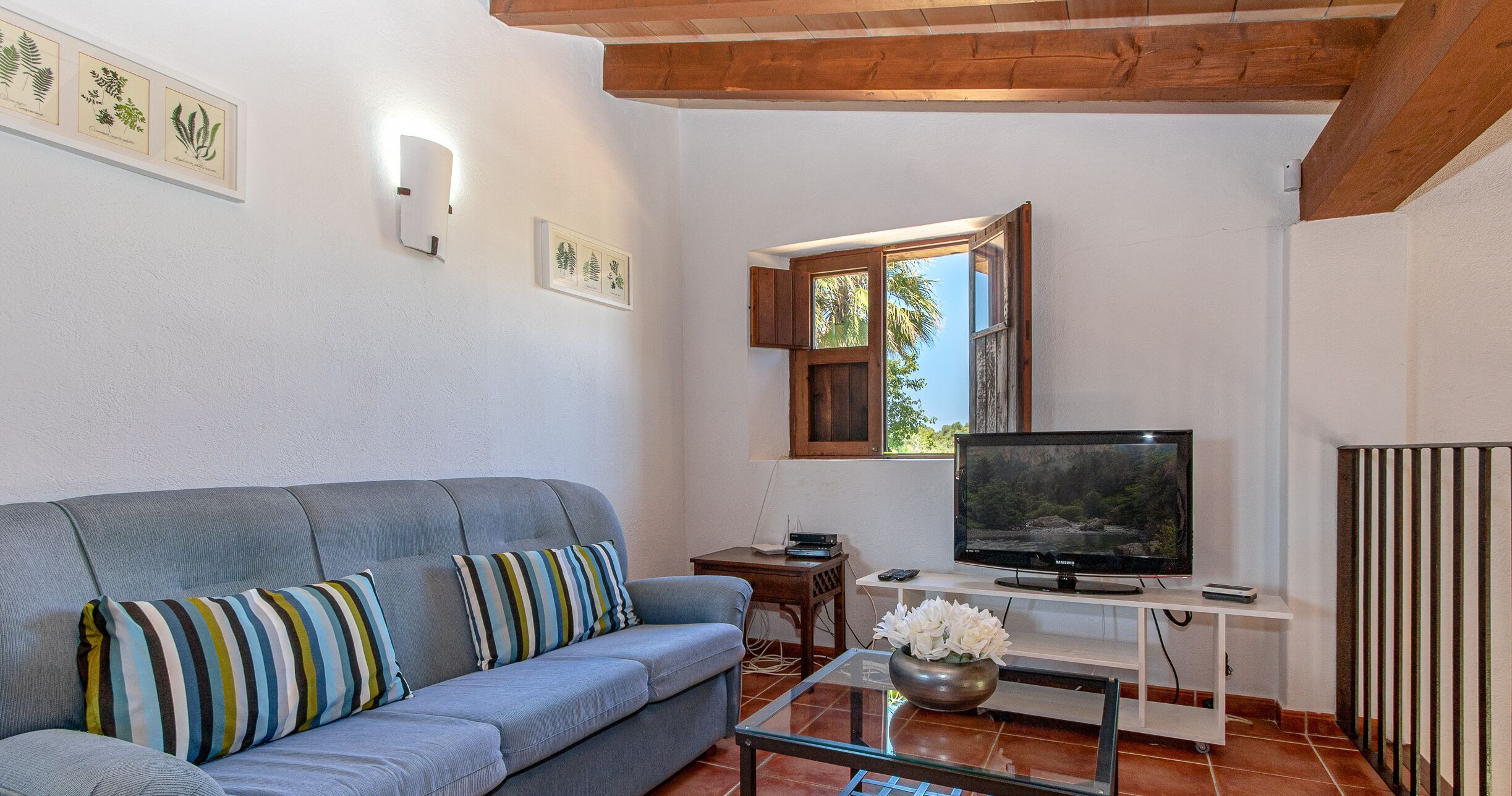 A living room with a sofa, a TV, a small table with a vase, and framed botanical prints on the wall.