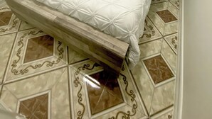 Una cama con una colcha blanca acolchada y almohadas, un cabecero de madera y una mesita de noche con una lámpara. El suelo tiene baldosas estampadas.