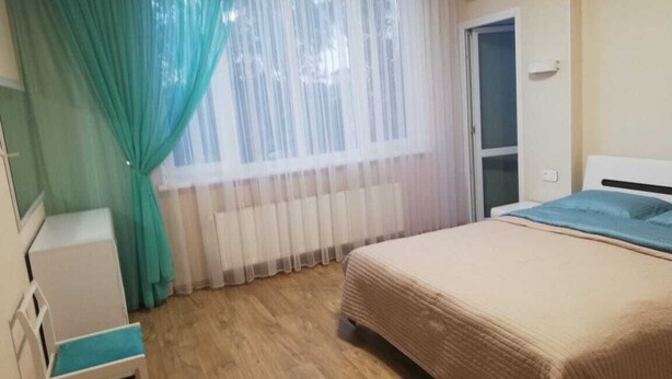A hotel room with a bed, a small table, a chair, a window with sheer curtains, and an air conditioning unit.