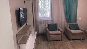 A modern living room with a flat-screen TV mounted on the wall, two beige armchairs with teal cushions, a wooden floor, and a window with sheer curtains.