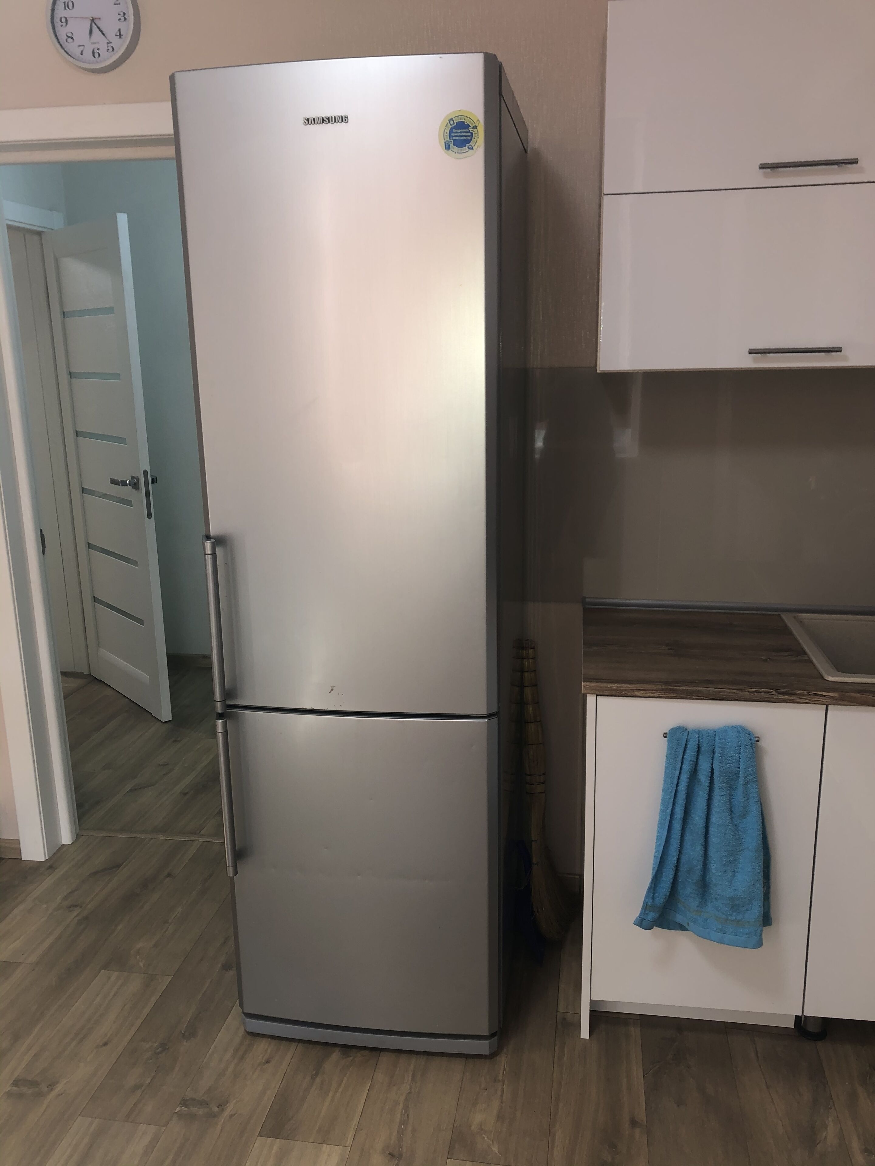 A stainless steel Samsung refrigerator in a kitchen with wooden flooring, a blue towel hanging on the cabinet, and a clock on the wall.