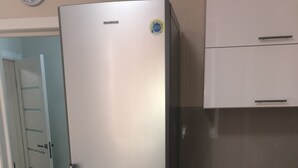 A stainless steel Samsung refrigerator in a kitchen with wooden flooring, a blue towel hanging on the cabinet, and a clock on the wall.