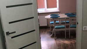 A room with a wooden floor, a white door with horizontal lines, a window with blinds, a table with blue cushions, and a chair.