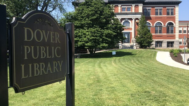 Une enseigne indiquant l’emplacement de la bibliothèque publique de Dover, devant un grand bâtiment en briques avec une pelouse.