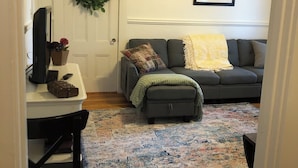 Un salon avec un canapé gris, une petite table sur laquelle se trouve un téléviseur, une couronne décorative et un ventilateur de plafond.