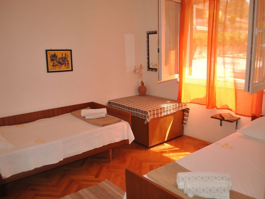 Une chambre avec deux lits simples, une petite table, un banc, un miroir et une fenêtre avec des rideaux.