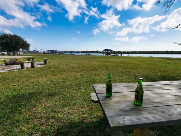 A grassy area with a wooden table and two green bottles, overlooking a body of water and a pier.