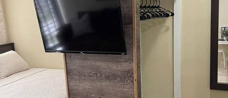 A room with a bed, a mounted flat-screen TV, a wooden panel, a glass shelf, and a power outlet.