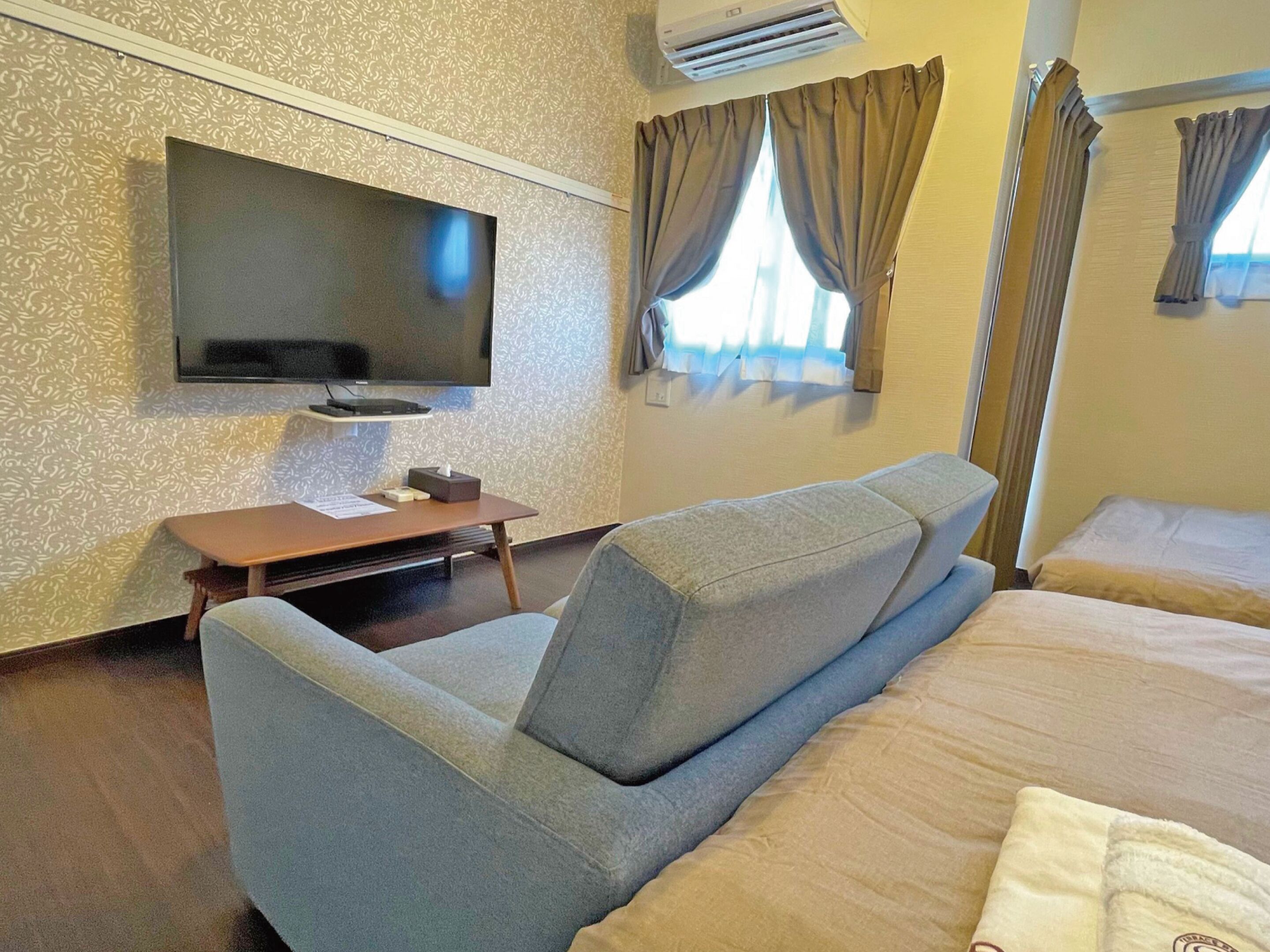 A hotel room with a flat-screen TV mounted on the wall, a sofa, a small table with a tissue box, and a bed with a white blanket.