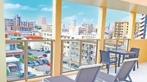 Un balcón con dos sillones de descanso y una mesa, con vista a un paisaje urbano que incluye varios edificios y coches estacionados.